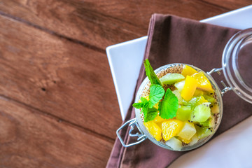 Healthy layered fruit dessert in glass jar. Kiwi, mango, pineapple, peach, chia seeds, mascarpone and whipped cream, mint and granola on brown wooden table. Top view. flat lay. Sweet menu concept.