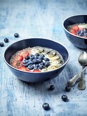 Two bowls of yoghurt and fruits - strawberries, blueberries and banana on blue table.