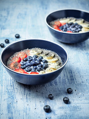 Two bowls of yoghurt and fruits - strawberries, blueberries and banana on blue table.