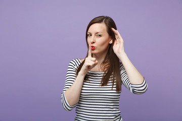 Secret young woman in striped clothes posing isolated on violet purple background. People lifestyle concept. Mock up copy space. Saying hush be quiet with finger on lips shhh gesture, try to hear you.