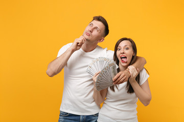 Pensive young couple friends guy girl in white blank t-shirts posing isolated on yellow orange background. People lifestyle concept. Mock up copy space. Holding fan of cash money in dollar banknotes.