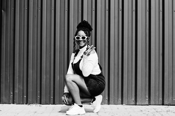 African woman in denim overalls skirt, white sunglasses crouching and show two fingers against green steel wall.