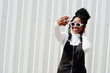 African woman in denim overalls skirt, white sunglasses posed against white steel wall and making selfie by phone.