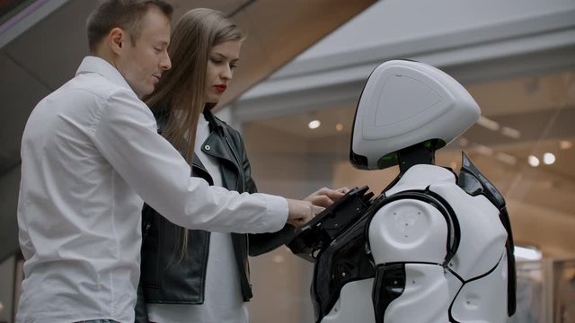 A man and a woman in the Mall interact with a robot consultant by tapping the screen and smiling. Cyborg Android helps people.