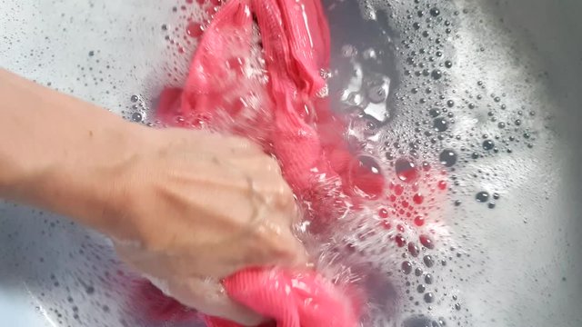 Female hand with blue nails wash a pink T-shirt in a basin, washing a dirty shirt with detergent, motion 4k footage close-up