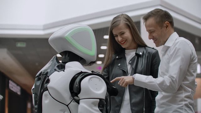 A man and a woman in the Mall interact with a robot consultant by tapping the screen and smiling. Cyborg Android helps people.