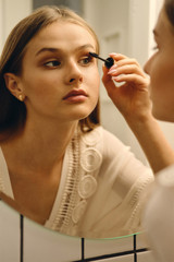 Young gorgeous woman in white dress and earrings standing near mirror and dreamily applying mascara in bathroom