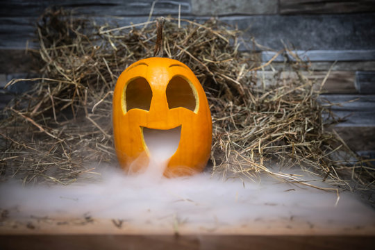 Yellow and very funny happy pumpkin for halloween celebration with smoke or vapor from the mouth. Stands on a wooden stand against a stone wall