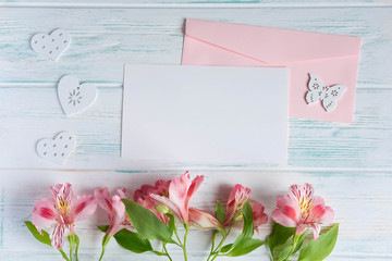 Mock up blank paper and envelope on white wooden background with natural flowers of pink color. Blank, frame for text. Greeting card design with flowers. Alstroemeria on wooden background.