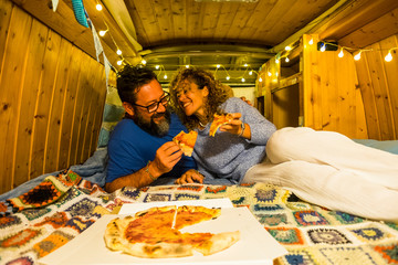 Happy couple of alternative traveler enjoying a pizza iside an old restored hand made vintage camper van - travel and relationship concept with nice people having fun. together