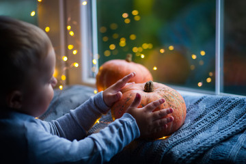 Toddler boy holding orange pumpkin on gray knitted plaid near window in evening surrounded with warm garland lights with golden bokeh. - 283834268