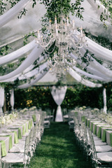 Outdoor summer wedding tent decorated with hanging fabric, greenery, and crystal chandeliers, wedding reception tables, green accent color