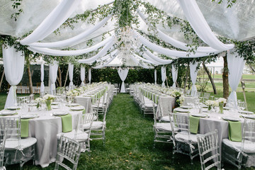 Outdoor summer wedding tent decorated with hanging fabric, greenery, and crystal chandeliers, wedding reception tables, green accent color