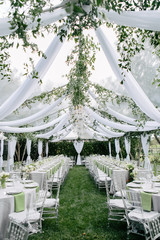 Outdoor summer wedding tent decorated with hanging fabric, greenery, and crystal chandeliers, wedding reception tables, green accent color