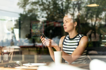 Thoughtful caucasian woman holding mobile phone while looking through the coffee shop window during...