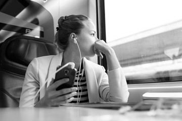 Thoughtful businesswoman looking trough the window, listening to podcast on cellphone using...