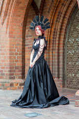 Fototapeta na wymiar Portrait of Tranquil Gothic Girl in Long Black Dress. Wearing Artistic Feather Crown with Roses. Posing Against Old Castle Gates.