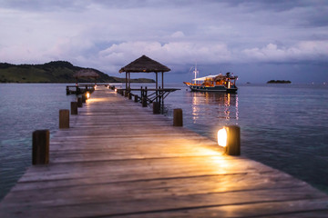 View of tropical island, pier and yacht tourist boat standing overnight in twilight with evening lights in bokeh. Marine travel and vacations concept.