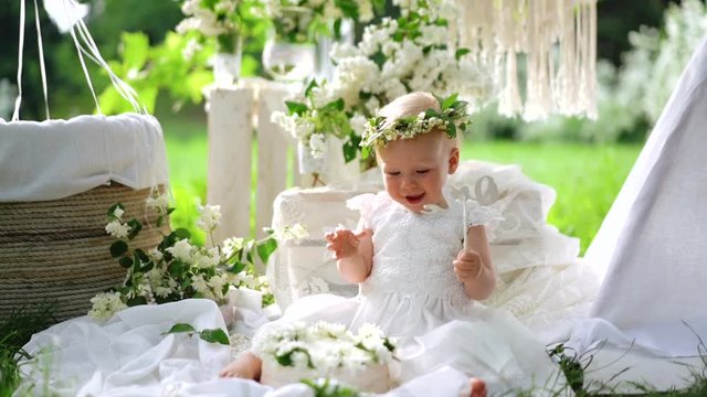 A little baby in the white dress and flowered wreath sits on the decoration and smiles.