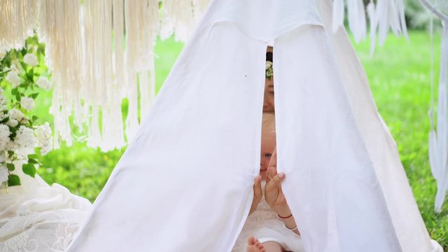 Mother with baby girl is having fun at celebrating birthday. A little girl with her mother hides in the white tent in the park. Portrait. Party atmosphere.