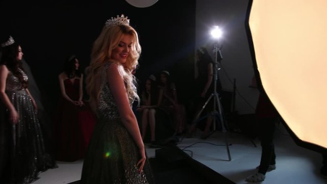 blonde model in a shiny lush dress, with a naked back, with a beautiful hairstyle and bright makeup, with a crown on her head, posing smiling, on a black background, photo shoot in Studio, slow motion
