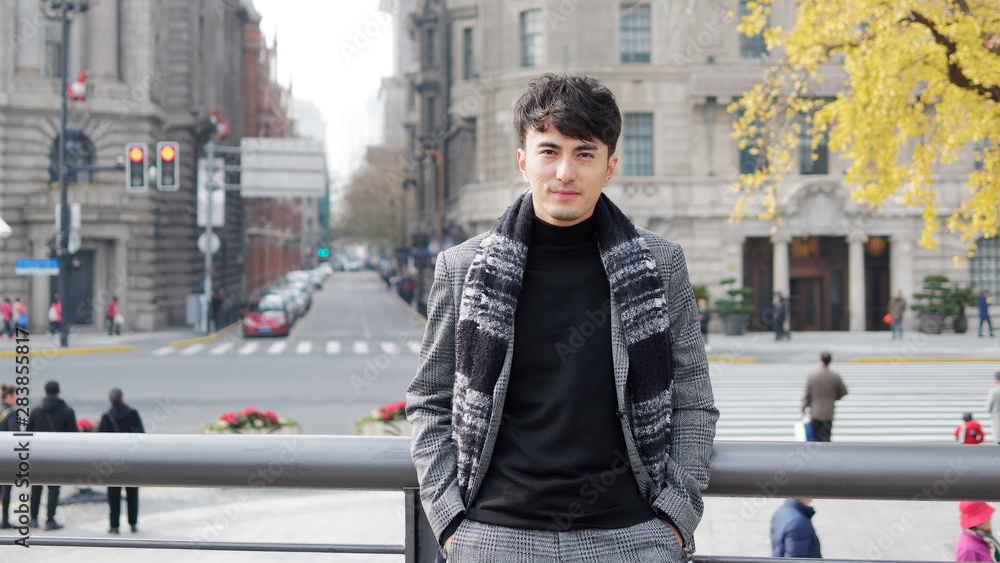 Wall mural Portrait of a handsome Chinese young man in casual suit smiling and looking at camera confidently with Shanghai bund background, winter fashion, cool young man lifestyle.