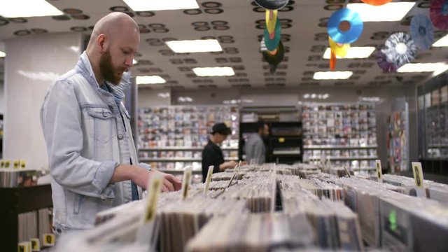 Thigh-up arc shot of young male hipster with shaved head and full beard, dressed in denim jacket, standing in record shop and looking through shelves of cds for sale, with other shoppers in background