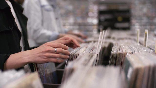 Trucking close-up hands shot of casually dressed unrecognizable female and male customers leisurely perusing cds and vinyl discs, grouped in alphabetical order, in small record shop