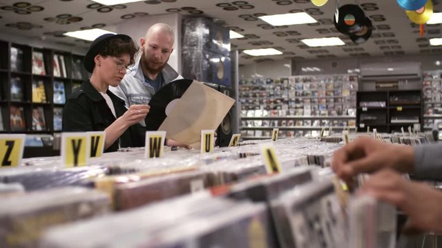 Trucking waist-up shot of happy stylish Caucasian couple choosing vinyls for purchase in small record shop, chatting and showing each other discs in plain white covers