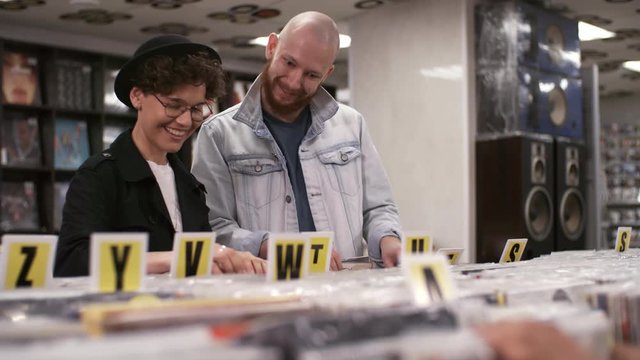 Waist-up shot of happy Caucasian girlfriend, wearing bowler hat and round glasses, and boyfriend with shaved head and beard, looking through selection of discs and admiring rare vinyls in record shop