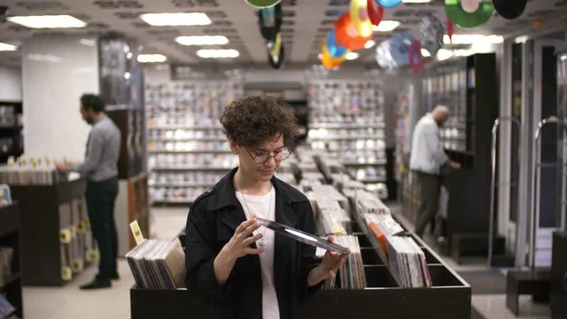Portrait shot of curly Caucasian woman in mid-thirties standing in record shop premises and checking out both sides of vinyl record for scratches, then looking up at camera with surprise and smiling