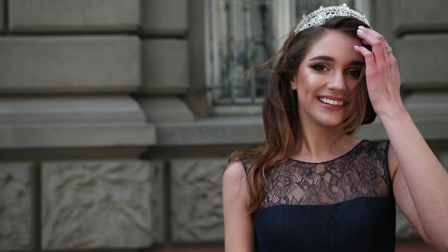 charming girl, brunette, in a dark blue long dress with lace, with curls and bright makeup, crown on her head, posing and smiling in front of the camera. is happening on the street near buildings