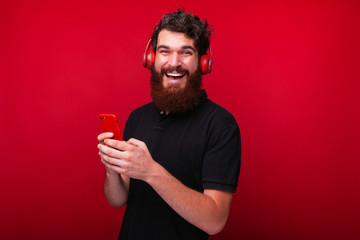 Handsome bearded guy, using a smartphone, and using headphones, looking at camera, standing over red background