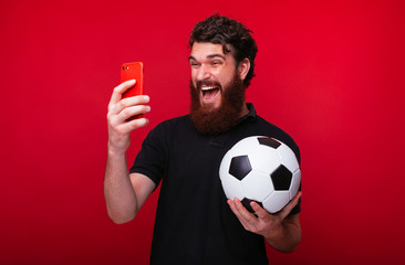Photo of bearded guy, holding a soccer ball, screaming and watching game online, over red background