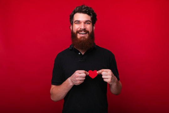 Love concept, handsome bearded guy holding a little paper heart over chest, looking at camera over red background