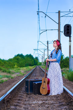 Beautiful girl with a guitar. Pretty woman with old suitcases on the train tracks. A teenager in a denim jacket, a long dress and a red bandage.