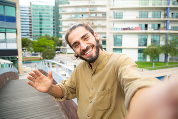 Joyful happy hipster guy waving hello at phone camera. Young man in casual using smartphone for...