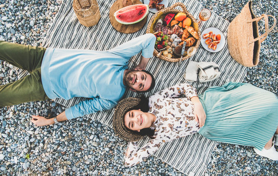 Summer beach picnic at sunset. Young couple lying on striped blanket, looking above at weekend picnic outdoor at seaside with sparkling wine, fresh fruit and tray of tasty appetizers, top view
