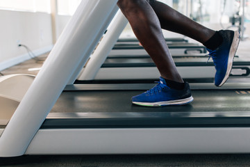 Side view of African American sportsman in blue sneakers working out on electric treadmill in fitness club. Faceless male runner running on treadmill