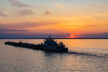 Obraz premium A tug on the river pushes a barge with sand or gravel. Sunset on the Volga river in Russia in July. Orange sun in the sky. Picturesque nature. Summer evening. River cruise