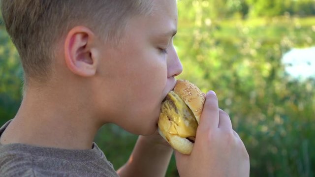 Closeup view of hungry kid eating big double cheeseburger standing outdoor. Slow motion full hd video footage.
