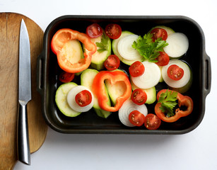 Vegetables in  black baking dish, bell pepper, zucchini, onion, cherry tomatoes and parsley, wooden board, knife, cut vegetables, cooking