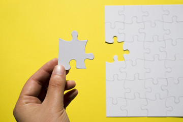 A person's hand holding missing white jigsaw puzzle piece over yellow backdrop