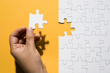 Human hand holding puzzle piece over white puzzle grid over yellow backdrop