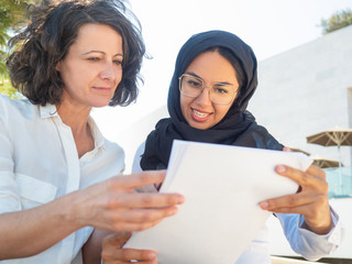 Positive focused female coworkers reviewing paper reports. Multicultural businesswomen in office clothing and Arabian veil reading and discussing document outside. Business documents concept