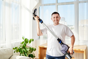 Portrait of young man in white shirt and jeans holding vacuum cleaner in living room or office, copy space. Housework, cleaning and chores concept
