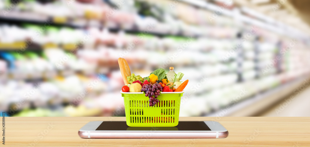 Wall mural Fresh food and vegetables in shopping basket on mobile smartphone on wood table with supermarket aisle blurred background grocery online concept