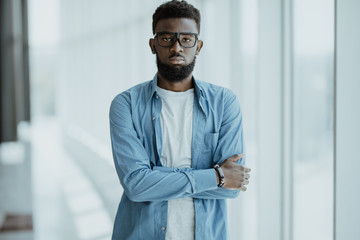 Thoughtful young african businessman with arms crossed in office against panoramic window