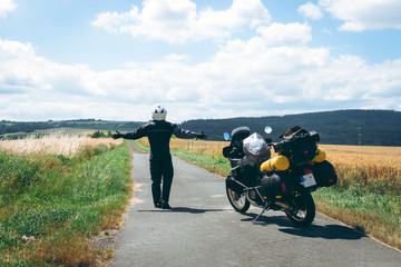 Obraz premium A male driver in a raincoat is standing by adventure motorbike his arms outstretched. a motorcycle tour journey. Outdoor, field and sky. World travel on two wheels, freedom concept. rider equipment.