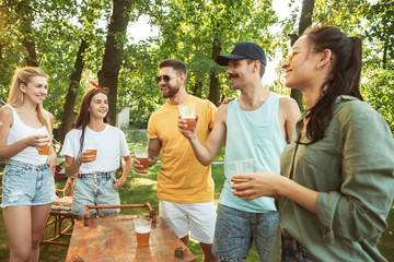 Group of happy friends having beer and barbecue party at sunny day. Resting together outdoor in a forest glade or backyard. Celebrating and relaxing, laughting. Summer lifestyle, friendship concept.